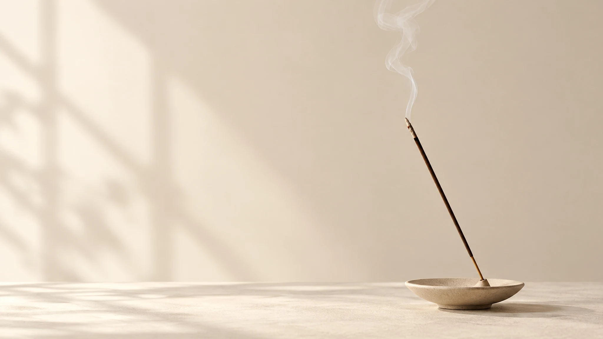 A lit incense stick leaning in a stoneware dish, window light falling across a pale wall
